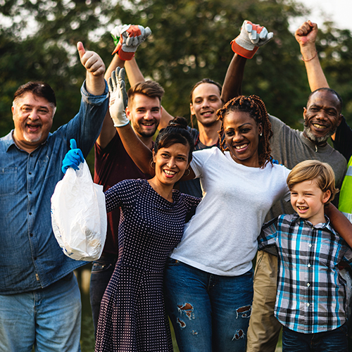 A group of people of different ages, races, and genders raise their hands in shared celebration as they perform cleanup in a park environment.