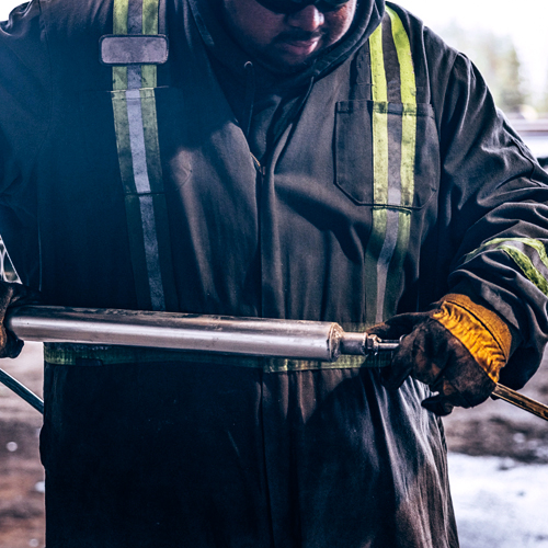 A worker in overalls and sunglasses concentrates on a cylindrical steel tool he holds in both hands.