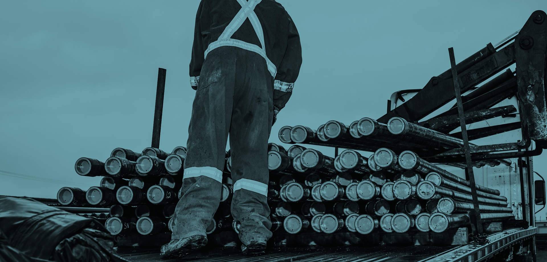 A worker in worn and rugged overalls has their back to camera as they inspect a rack of approximately 50 heavy-duty oilfield pipes.