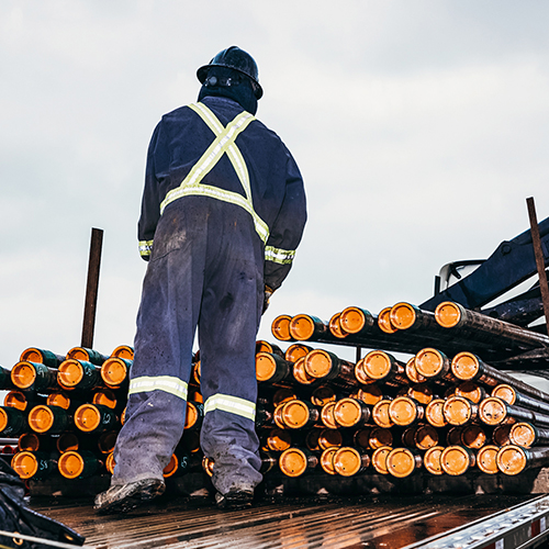 A worker in worn and rugged overalls has their back to camera as they inspect a rack of approximately 50 heavy-duty oilfield pipes.