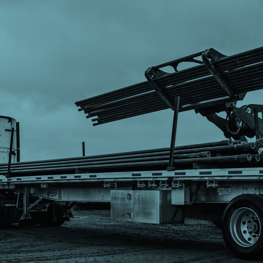 A worker wearing overalls squats in the bed of a large truck as he supervises a heavy load of oilfield piping being lowered onto the truck for transportation.