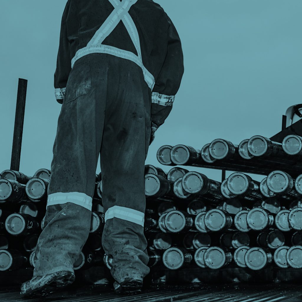 A worker in worn and rugged overalls has their back to camera as they inspect a rack of approximately 50 heavy-duty oilfield pipes.