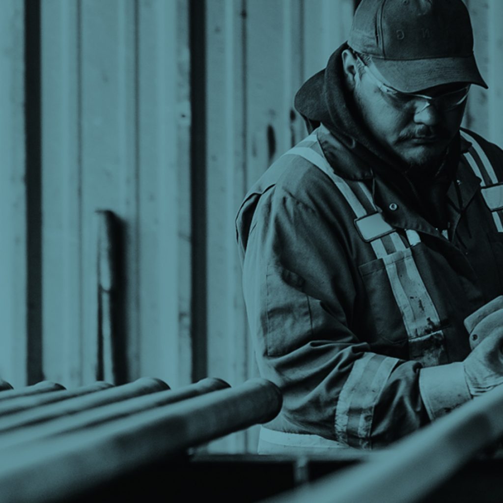 A worker in heavy overalls, gloves, protective eyewear, and a baseball cap prepares to perform a service upon a row of oilfield pipes with a specialized tool held in both hands.